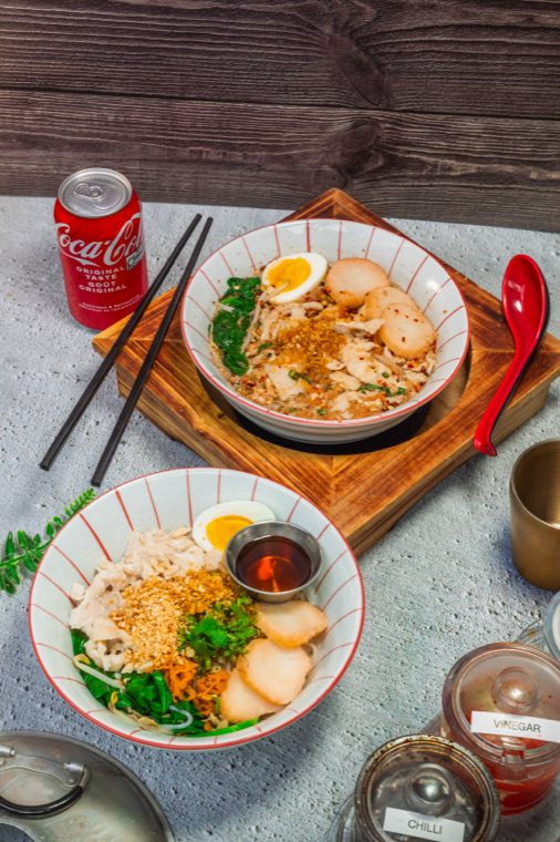 Two small noodle bowls on a dark wood table, each topped with a soft egg, greens and fried garlic