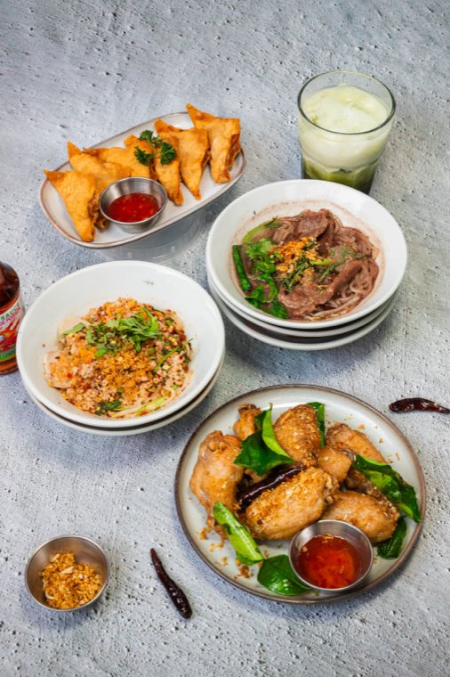 A shared table: fried chicken wings, spring rolls, fried rice and two stacked noodle bowls